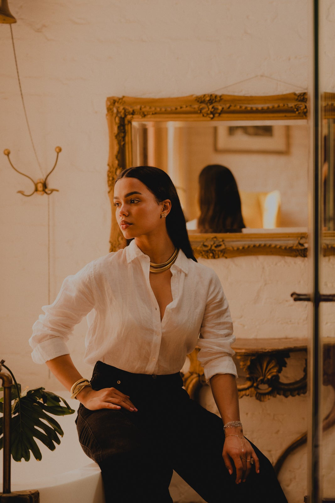 model in white linen blouse and black jeans sitting on the side of a claw foot tub with vintage mirror hanging on the wall. Model is wearing pieces from the Rumours Tobogas collection set in 14k and 18k gold. Bracelets and necklaces are tubogas style jewelry and rose cut diamond multi strange bracelets.