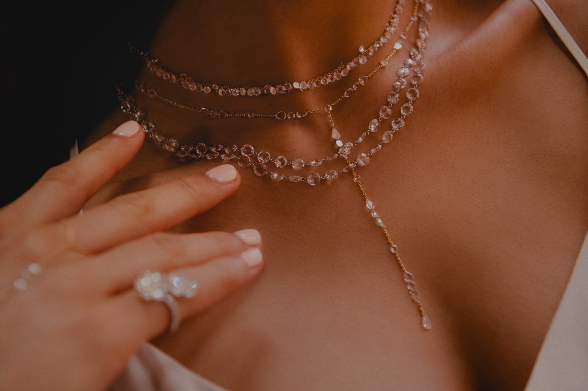 Woman's décolletage and hand wearing layered rose cut diamond jewelry. 4 necklaces layered and 1 lariat all sparkle in the light. pinky ring is out of focus which is flowers made out of rose cut diamonds and hand chain can be seen.