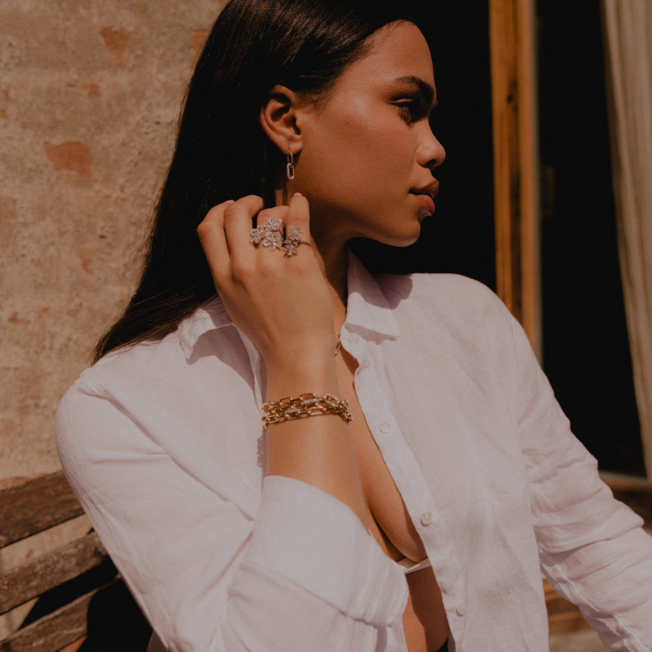 Model sitting outside, side profile showing 14k gold and diamond pave earrings. 2 Flower rose cut diamond rings and chain bracelet made out of 14k yellow gold and pave diamond links.