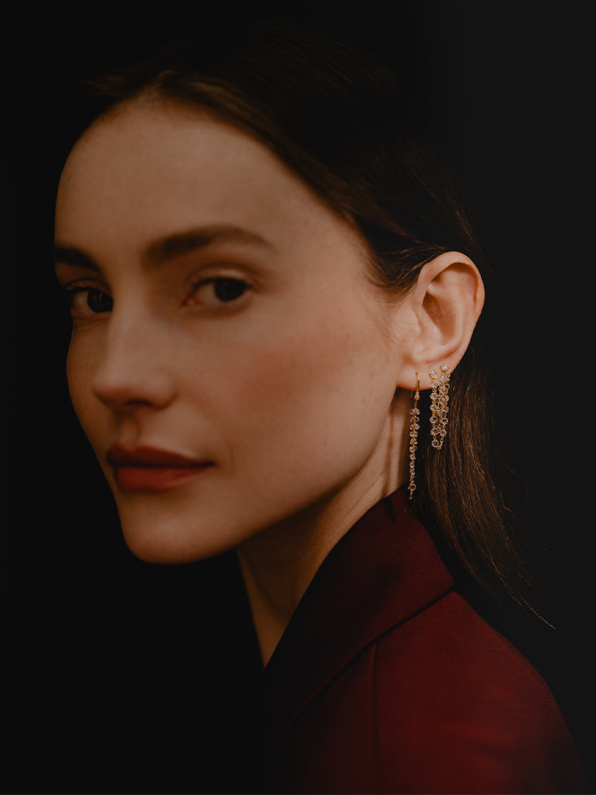Side profile or woman's face wearing dark burgendy blazer with hair pullled back so you can see her one ear which has 2 earrings of dangling rose cut diamonds and 18k yellow gold. They are drapping down for a cascading effect.