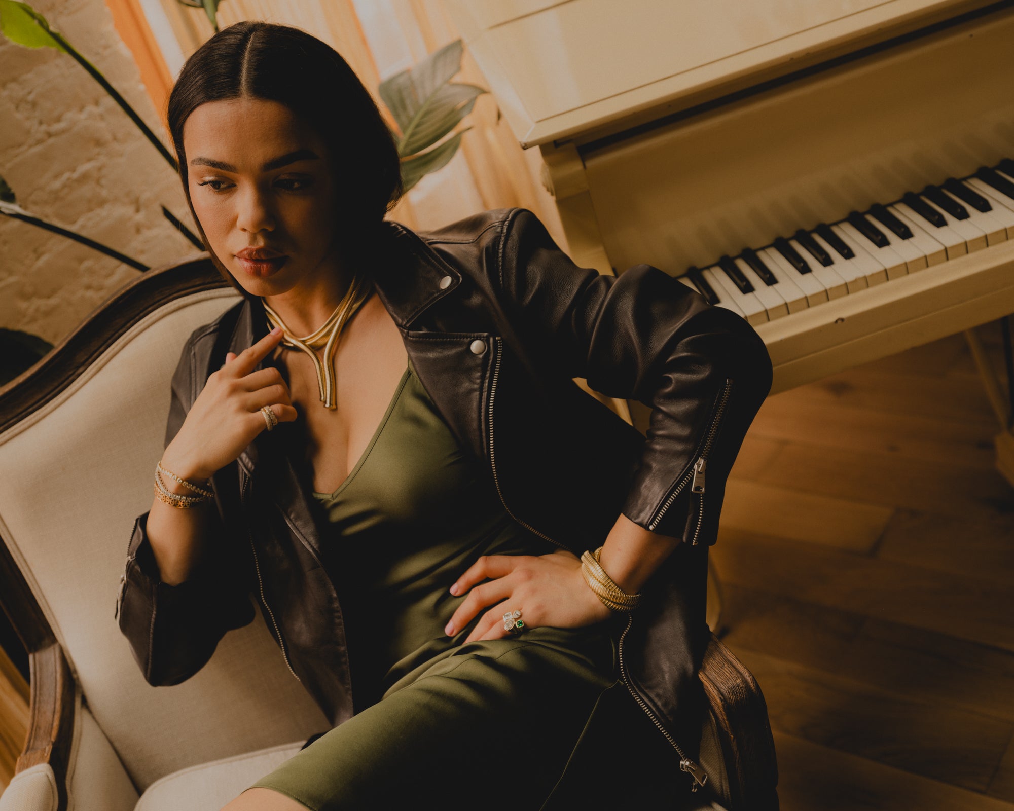 Woman in a green dress and black leather jacket sitting next to a piano wearing yellow gold tubogas laybered necklaces. 