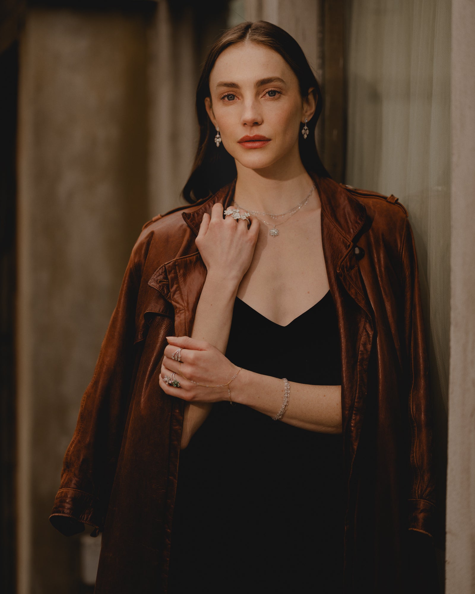Woman wearing a brown leather coat over a black dress, styled with diamond jewelry including floral rings, drop earrings, a delicate necklace, and a bracelet, standing against a softly lit urban backdrop.