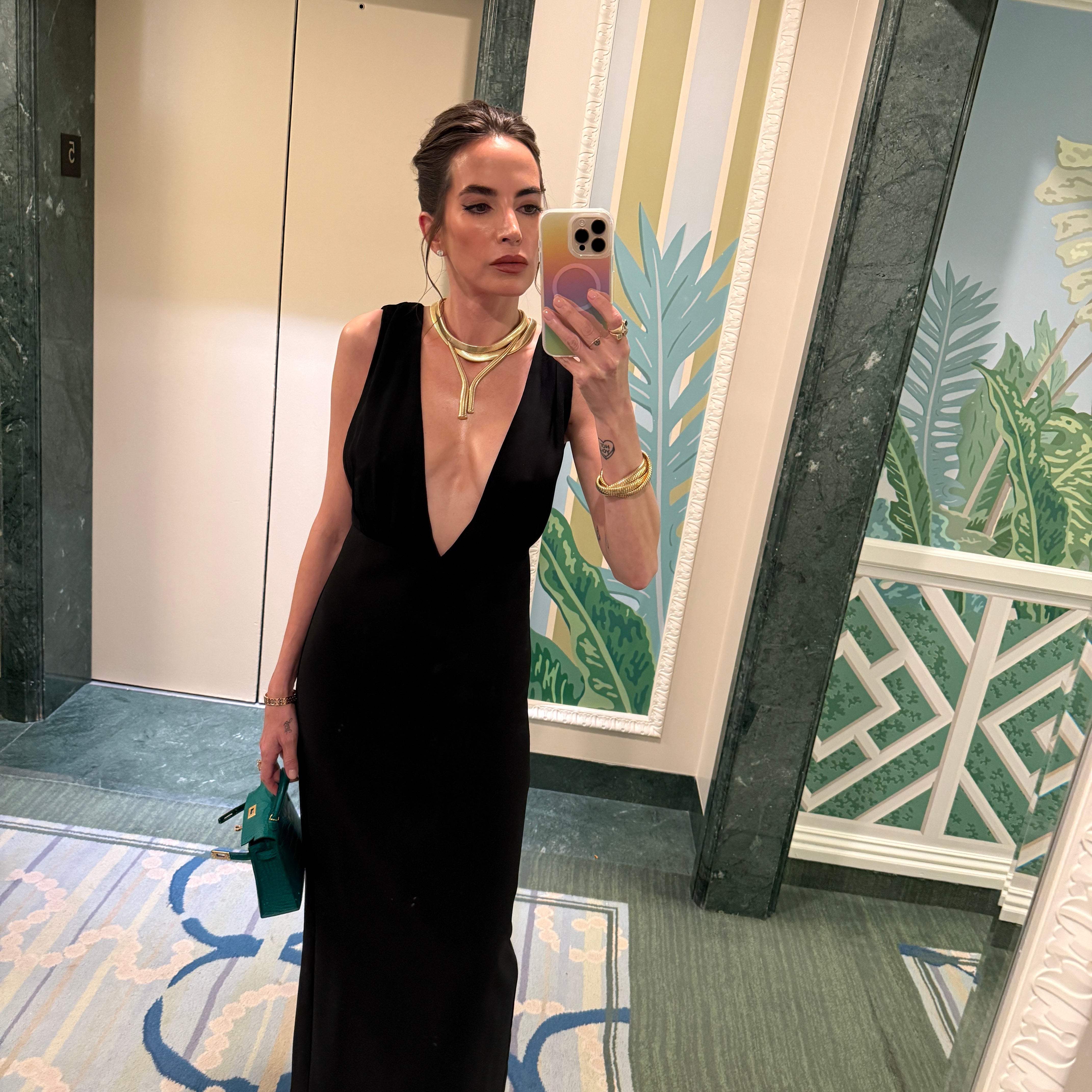 Person wearing a black dress standing in front of a decorative mirror with floral patterns. Katie Mancilla wearing a black Reformation dress, 18k gold tubogas necklaces and bracelets.  Carrying a green Hermes Mini Kelly.