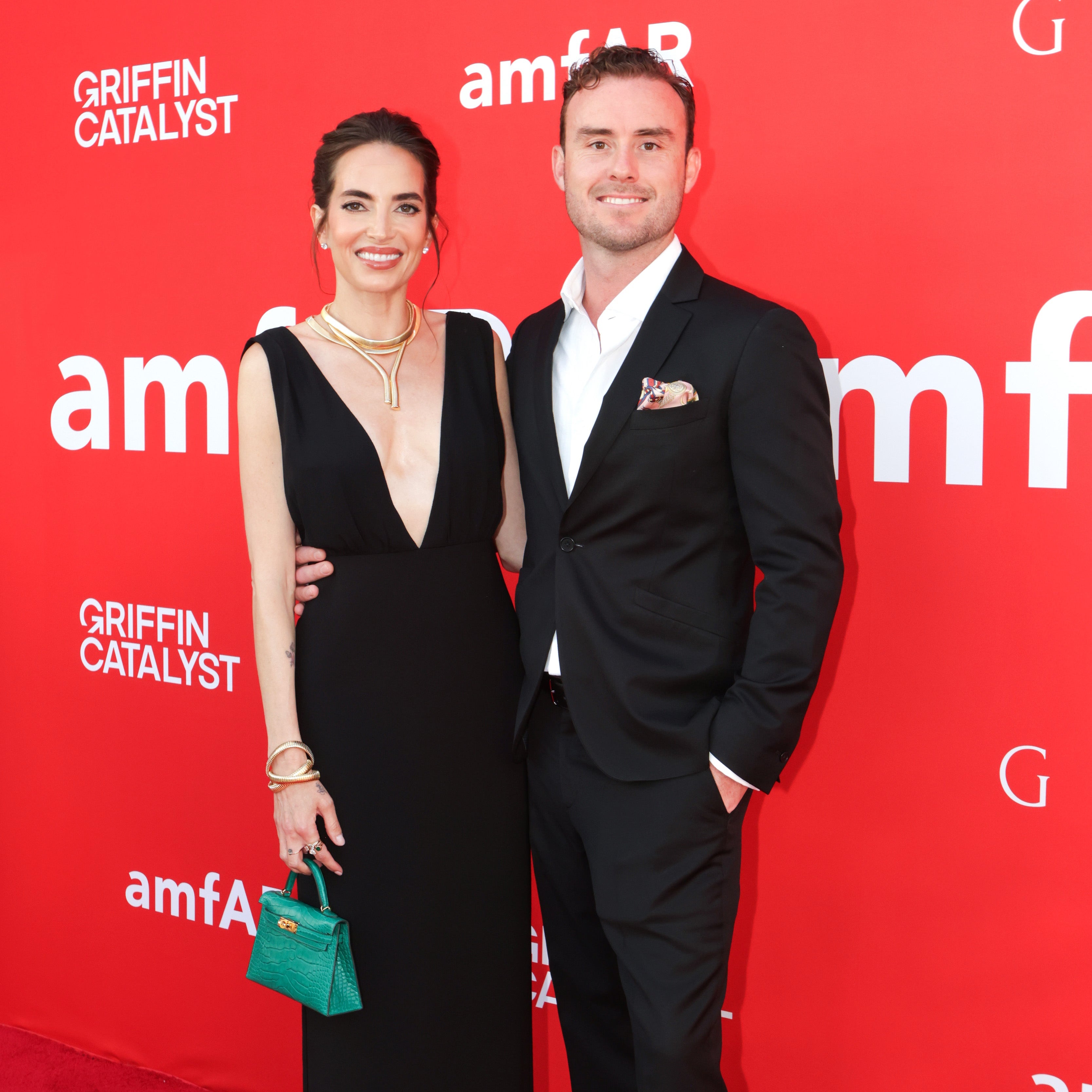 Man and woman posing on a red carpet event with 'amfAR' branding. Woman Katie Mancilla wearing a black Reformation Dress and Gold Tubogas Necklaces and Bracelets and carrying a green Hermes Mini Kelly.  Man Tony Mancilla wearing a black suit. 